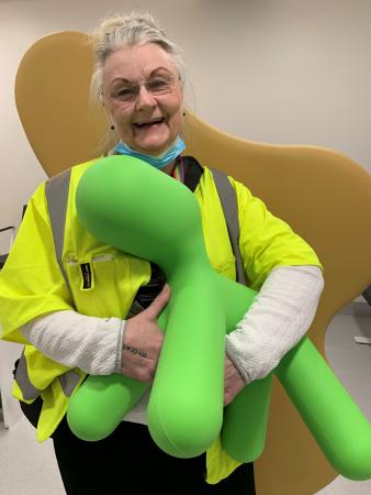 Agneta har grått uppsatt hår. Hon bär en gul väst och håller i en ljusgrön hundliknande leksak som man kan sitta på i väntrummet, akuten.