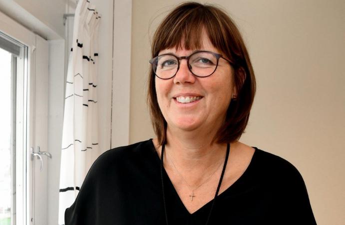 Pia har mörkt, axellångt hår och glasögon. Hon bär en svart tröja och skrattar på bilden taget bredvid ett fönster.