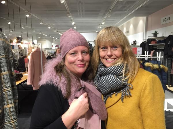 Eva Marie Svensson står till vänster i bilden iförd den rosa pälskrage som Ann-Sofie talar om i reportaget. Hon har matchande mössa och vantar i ull. Hon ser nöjd ut. Ann-Sofie bär en gul ullkappa som hon beskrivit i reportaget. Till vänster skymtar man en annan omnämnd kappa i grått med rutor i vitt, svart och gult. Båda står mitt i varuhuset Åhlens i Luleå omgivna av kläder.