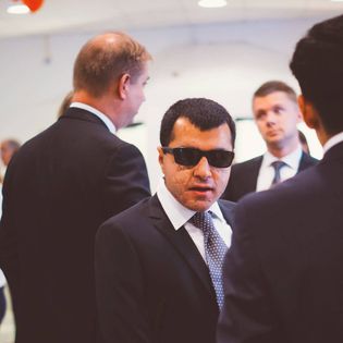 Ther har svart kostym och vit skjorta med slips. Han har mörkt kort hår och solglasögon. Han står omgiven av andra män i kostym.