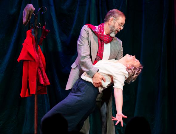 Tomas iklädd kavaj med en röd sjal slängd över axeln håller en nedböjd Karin i en dramatisk dansposé.