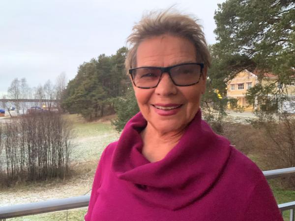 Ulla har kort, brunt hår och glasögon med mörk båge. Hon bär en plommonlila topp med vid tubkrage.