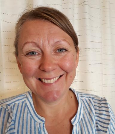 Pia har uppsatt mörkblont hår och ler på denna porträttbild. Hon har en vit och ljusblårandig skjorta.