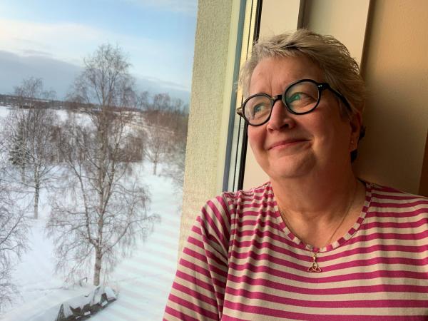 Gunilla har ljusT kort hår och bär glasögon med mörk båge. Hon bär en röd- och vitrandig tröja och ser glad ut. Hon tittar ut genom ett fönster.