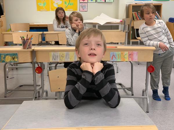 Viktor sitter i sin bänk i klassrummet. Han lutar hakan mot händerna. Han har ljust hår och ser glad ut. Bakom viktor står och sitter tre klasskamrater