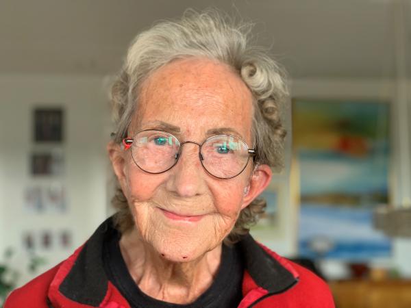 Elisabet har kort, grått, lockigt hår och glasögon. Hon bär en röd tröja med svart krage och ser glad ut.