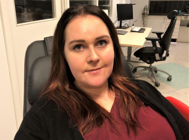 Sanna sitter i en stol och har långt mörkt hår. Hon ser glad ut och bär en röd tröja.