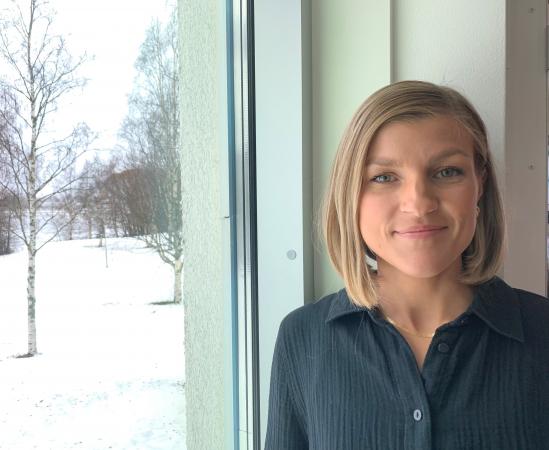 Johanna har en axellång page. Hon har ljust hår och en svart skjorta. Hon står framför ett fönster. Ute är det snö.