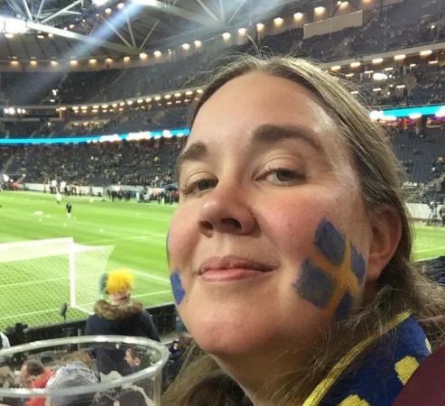 Sara sitter i publiken på en fotbollsarena och har svenska flaggan målad på kinden. Hon ser nöjd ut.