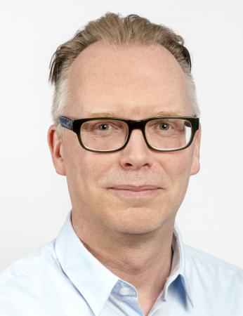 John har kort bakåtkammat hår och glasögon med mörkbåge. Han bär en ljusblå skjorta oc ser glad ut.