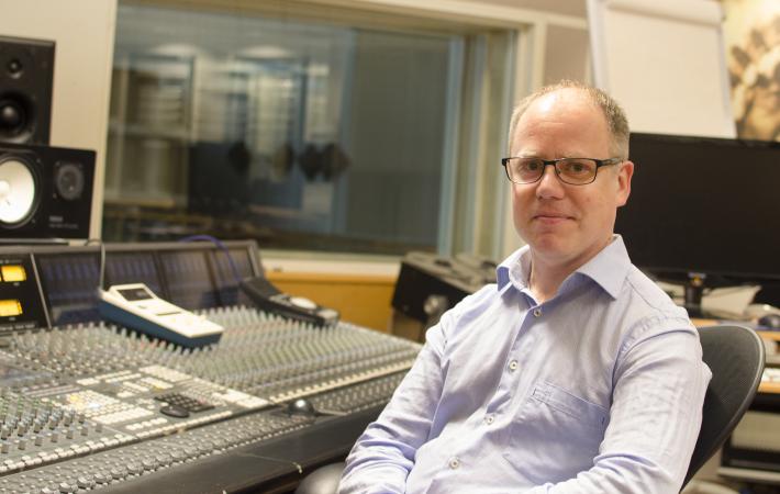 Jon Allan är snaggad och bär glasögon. Han har en skjorta på sig och sitter i en ljudstudio framför ett mixerbord. Foto: LTU