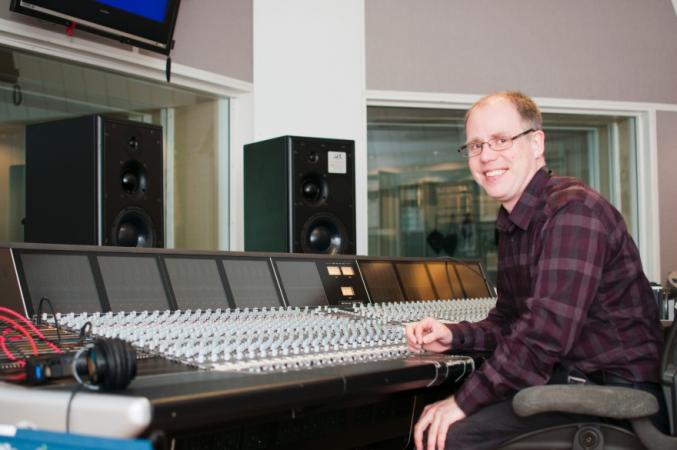 Jon sitter i en ljudstudio framför ett stort mixerbord. Han har kort brunt hår och bär glasögon. Han ser glad ut.