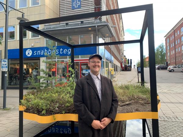 Andreas har svart kepa, glasögon och en svart kavaj. Han står på Storgatan i Luleå framför en ihopsvetsad ram i form av en kub. Han har hämtat växter från SSAB området och skapat verket Svartön. 
