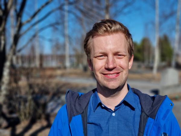 Anders står utomhus framför ett träd. Han har ljusbrunt kort hår, har blå jacka på sig och ler glatt.