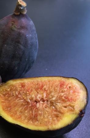 Ett halvt fikon ligger uppskuret på en mörk yta. Fruktköttet är brungult med många kärnor. I bakgrunden ett helt fikon med blålila skal.