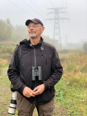 Christer står i en dimmig skog under en gigantisk kraftledning. Han har Kepa och bär glasögon. Detkortklippta håret är grått och runt halsen hänger en kikare och över axeln en systemkamera med ett gigantiskt objektiv.