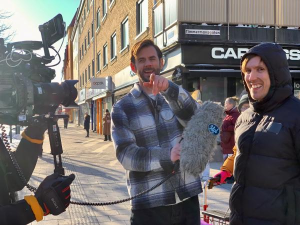 En filmkamera syns till vänster och Idols programledare pekar på taltidningens utsände. Han har en rutig skjorta. Luleåsångaren Niklas skrattar och har en svart täckjacka med huvan uppfälld. De står på Storgatan i Luleå.