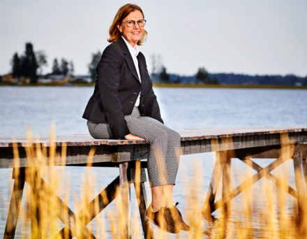 Anna-Stina sitter på bryggan på Fårö i Piteå skärgård. Hon har en mörk grå kavaj och ljusa byxor. Hon dinglar med benen och ser glad ut!