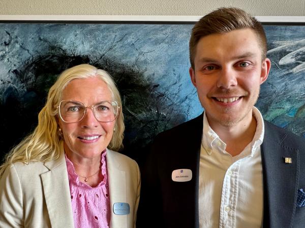 Ann Christine har långt blont hår och glasögon. Hon bär en rosa blus och en benfärgad kavaj. Jens har mörkt, kort hår och har en svart kavaj och vit skjorta. De står framför ett konstverk och ser glada ut.