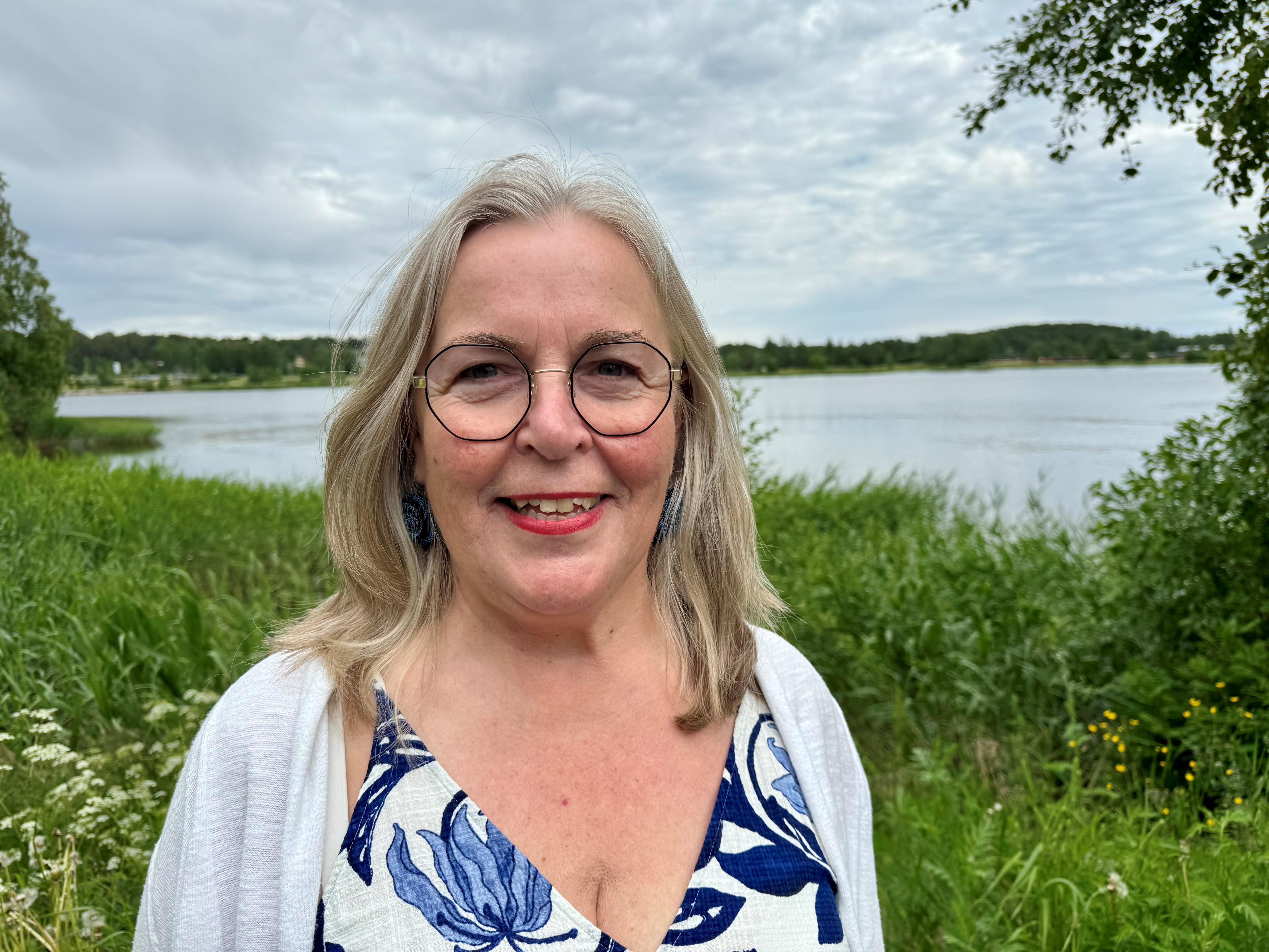 Anna-Maria är blond och har axellångt hår. Hon bär glasögon och ser glad ut. Hon har en klänning med blått mönster och en vit kofta. Hon står framför vatten och grönska