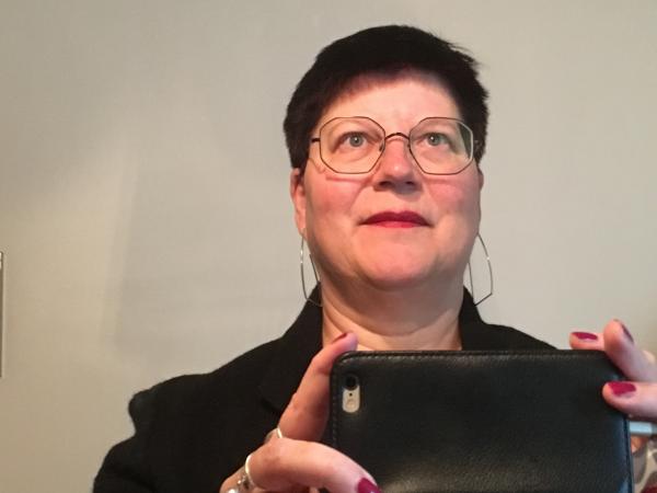 Susanne Jonsson tar en klassisk selfie med mobilen. Dvs hon fotar sig själv i en spegel. Något som många gör 2018. Susann har mörkt kortklippt hår. Hon bär glasögon och har rött läppstift. Fingrarna som håller i  mobilen är knallröda. Hon har en svart jacka. På bilden ser hon finurlig ut och blickar upp mot högra hörnet.