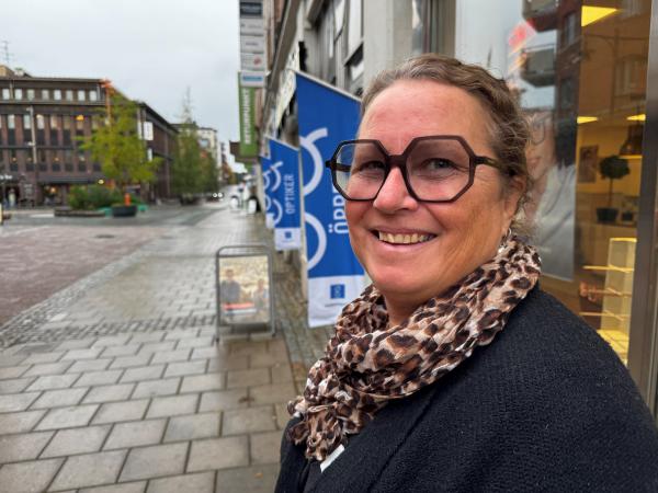 Karin har kantiga, stora, mörka glasögon i plast. Hon har det lockiga håret uppsatt och ser glad ut utanför glasögonbutiken