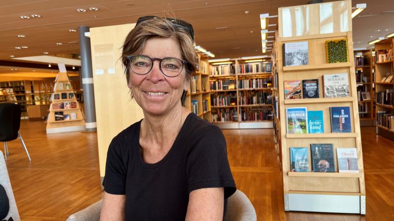 Marita har kort, mörkt hår och bär glasögon. Bilden är tagen på ett bibliotek och Marita ser glad ut.
