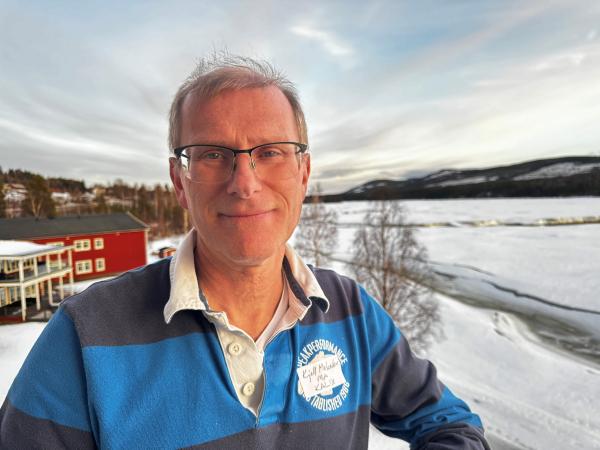 Kjell har kort hår och bär glasögon. Han har en blå randig tröja och står på en balkong med utsikt över Storforsen.
