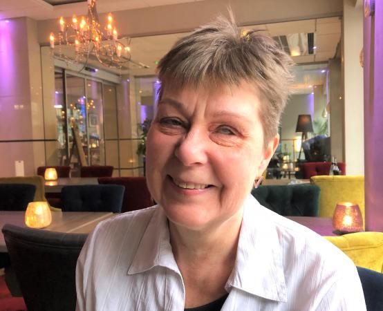 Karin Sundbaum har kortklippt, lite spretigt, ljusbrunt hår. Hon har en vit blus på sig och ler in i kameran. I bakgrunden syns bruna och gula möbler i hotell Savoys lobby. 