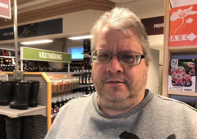 Mikael har ljust, kort hår och bär glasögon. Han har en grå tröja och står bland hyllor.
