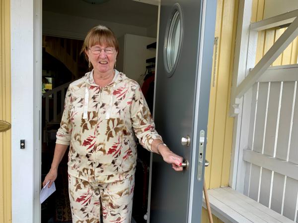 Monika tar emot Insyns reporter i dörren. Hon har en mönstrad, två delad dress och håret uppsatt. I handen håller hon kuvertet med de två trisslotter hon precis har fått och hon ser glad ut.s