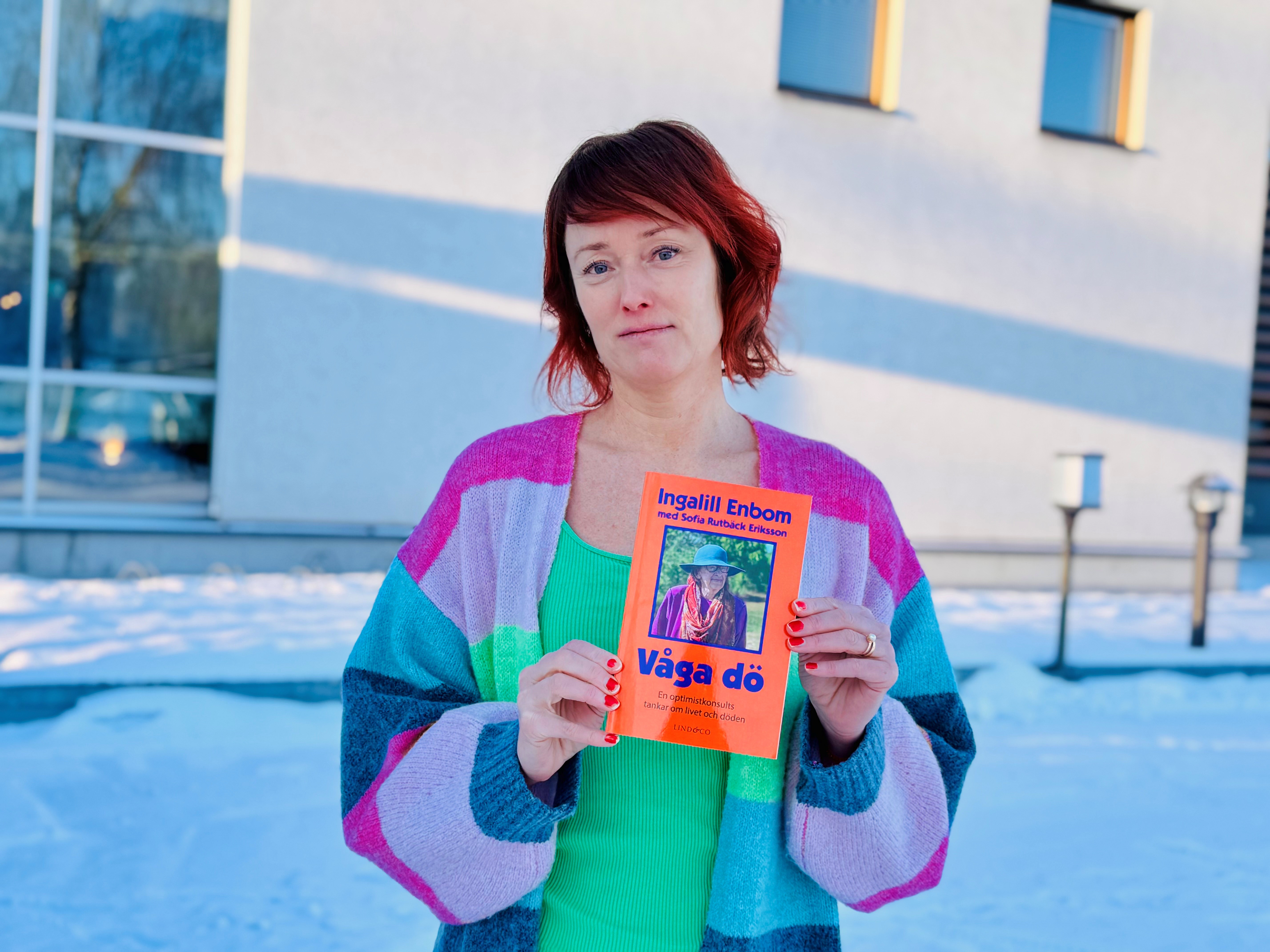 Sofia har rött, kort hår. Hon är iklädd en kofta i bland annat grått, rosa och blått. Under den har hon ett limegrönt linne. Hon håller upp en orange bok.