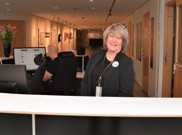 Anna-Greta har ljust hår och är klädd i svart. Hon står bakom servicedisken på Regionhuset i Norrbotten. Hon ser glad ut.