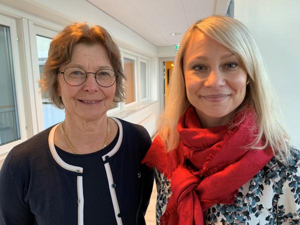 Eva klädd i svart har kort, brunt hår och glasögon. Helena är blond iförd röd sjal. Båda ser glada ut på bilden.