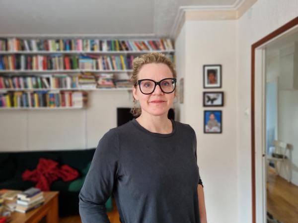 Johanna Hövenmark står i sitt vardagsrum framför en soffa och en bokhylla med massa böcker på. Hon har en grå tröja med långa ärmar. Håret är uppsatt och hon har svarta glasögon. Hon ler lite.