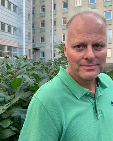 Lars står utanför sjukhuset i Umeå och har bär en grön piketröja. Han har grått, snaggat hår och ser glad ut på bilden.