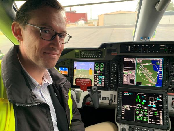 Mikael har kort mörkt hår och glasögon. Han sitter i cockpit på ambulansflyget och bakom honom ser man instrumentpanelen som är helt digital och består av många skärmar.