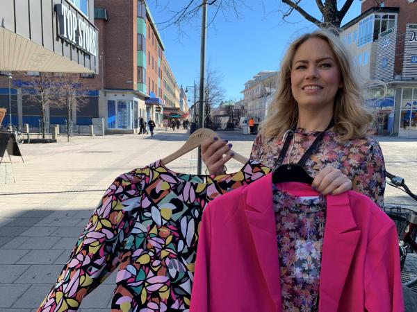Elisabeth har långt blont hår och blå ögon. Hon har på sig en småblommig blus och håller i en chockrosa kavaj och en småblommig blus i rosa och turkos.