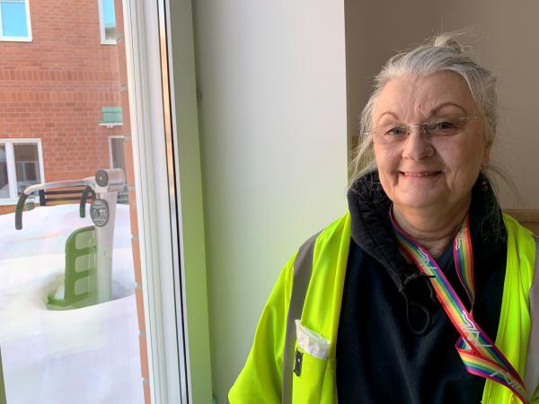 Agneta har grått, uppsatt hår och bär en gul reflexväst. Hon står framför ett fönster och man ser en utegård i bakgrunden 