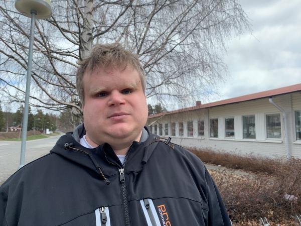 David har lockigt, ljust hår och står ute framför ett hus.