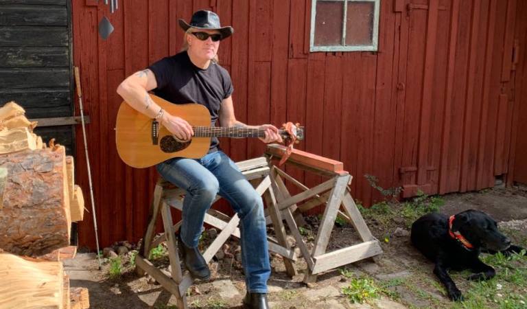 Johan sitter ute på en plint vid en husvägg ch en vedhög. Han har en svart t-shirt, blå jeans och en cowboyhatt. Han håller i en gitarr