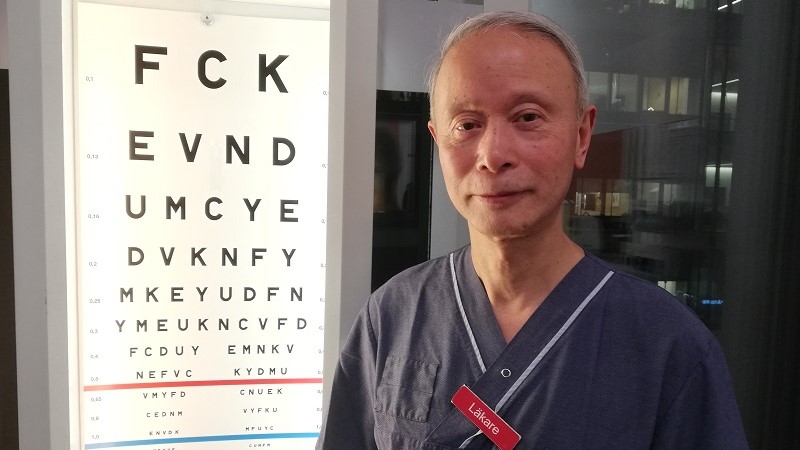 Enping står brevid en syntavla och har grått kort hår. Han är klädd i blå doktorsskjorta och har en röd namnskylt där det står läkare.