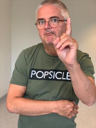 Anders ser klurig ut och hötter med vänster pekfinger. Han har grått kort hår, glasögon och bär en mossgrön t-shirt som det står popsicle på
