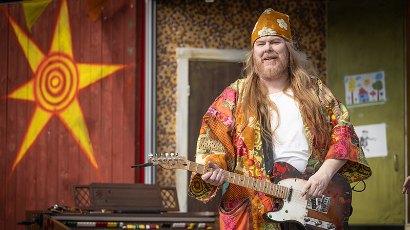 Mattias har en orange tehuva med moln på huvudet och en lapptäckeinspirerad kaftan. Han står på scenen och spelar gitarr. Han ser glad ut.