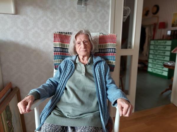 Birgitta Hansson sitter i en fåtölj. Hon har en blå kofta över en grön tröja. Hon ser allvarlig ut. 