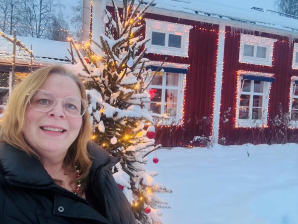 Eva Marie står ute i ett vintervitt Boden framför den röda norrbottensgården som kallas Norrigården. Eva Marie ser glad ut, har ljust hår och glasögon. Hon står framför en pyntad en julgran.