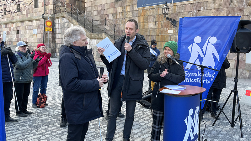 Jens står på Mynttorget utanför riksdagen i Stockholm och håller papper i händerna. Runtomkring står människor och applåderar. 