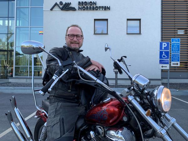 Roger står framför regionhuset och sitter på en långgafflad, röd motorcykel. Han har en svart skinjacka, håller hjälmen i knät och ser glad ut. Han har kort lockigt hår och glasögon med mörk båge.