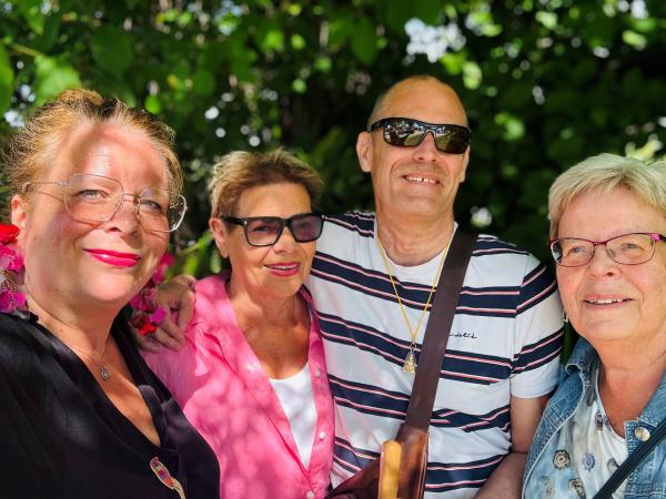 Eva Marie har uppsatt hår och rosa dinglande örhängen med matchande läppstift, Ulla Bergeros har en barbierosa blus och är korthårig, blond med mörka partier, Mats har en svart och vit randig t-shirt och bär solglasögon. Han har inget hår. Marja Liisa är blond och korthårig, med glasögon och en ljusblå blus. Alla är glada!