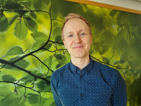 Marcus Östling har en blå skjorta på sig. Han är blond och står framför en vägg med en grön tapet med mönster av trädgrenar.